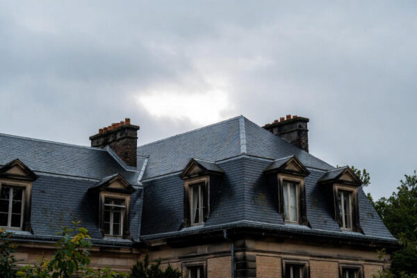 A classic mansard roof with slate tiles and dormer windows on a traditional residential building under an overcast sky.
