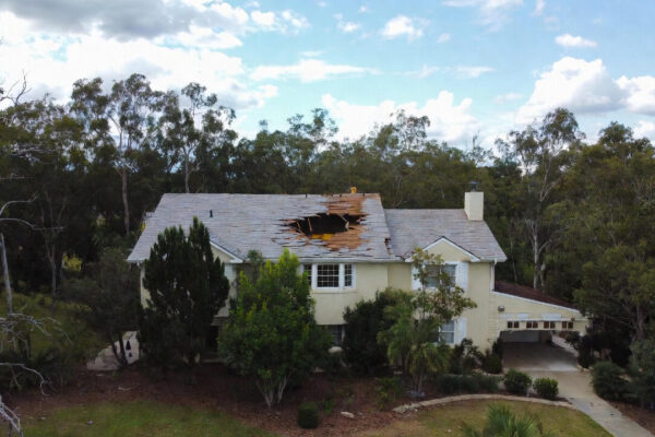 A residential home with severe storm damage to a gable roof, showing broken shingles and a large hole requiring urgent roof repair.